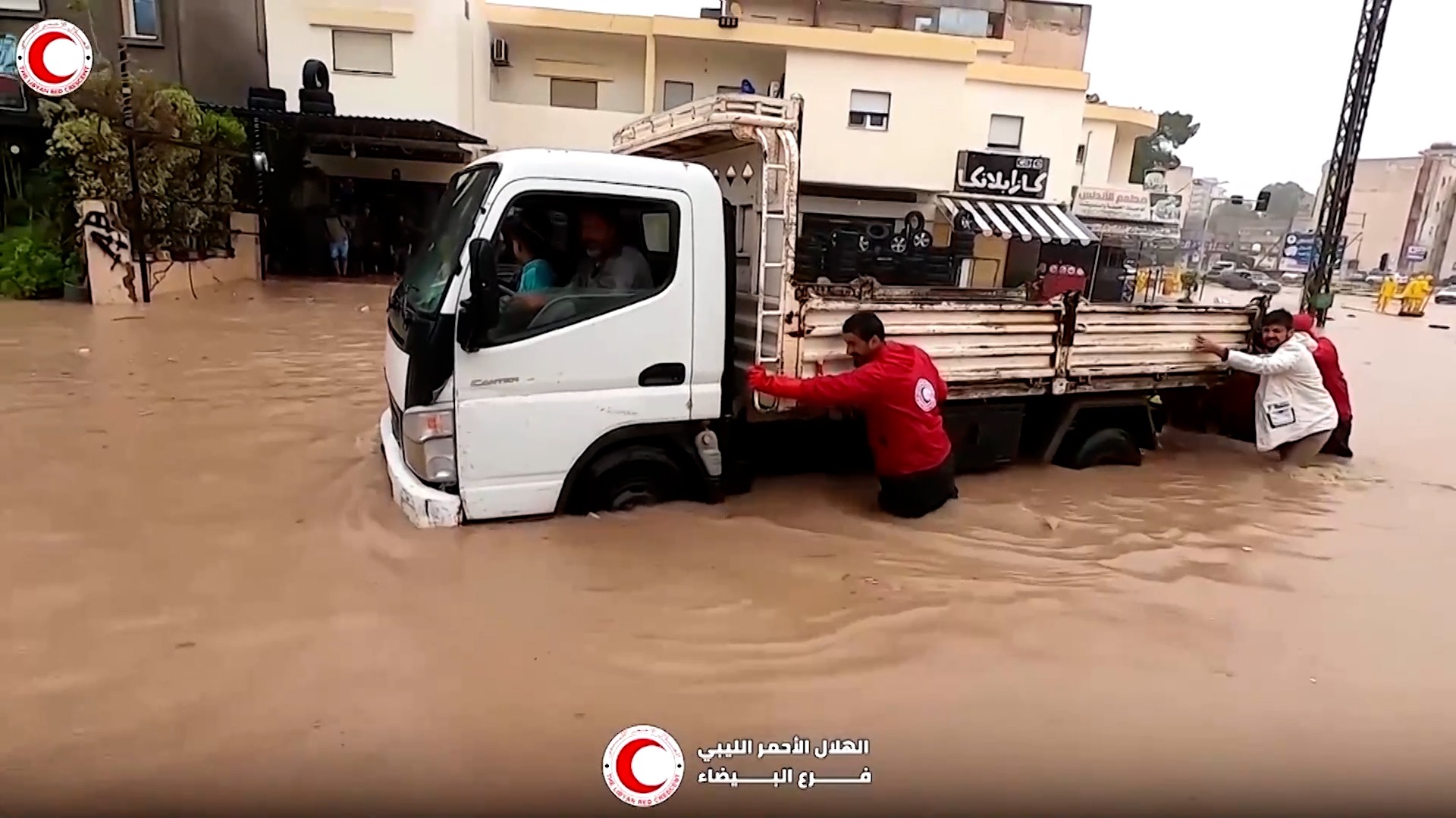 Red Crescent workers helping motorists through flooded streets. The Red Crescent workers push a small builders truck through brown floodwater that almost covers the vehicle’s tyres