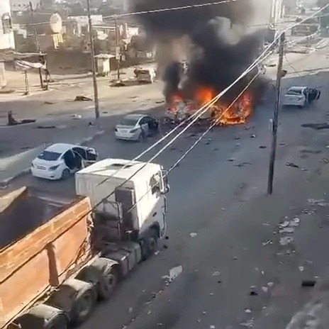 Smoke billows from two flaming cars in the middle of a wide road. Other cars sit behind them, abandoned, as people run away